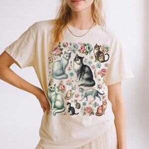 May include: A cream-colored short-sleeve t-shirt featuring a graphic print of multiple cats in various poses interspersed with colorful floral illustrations including pink and blue flowers across the front of the garment.