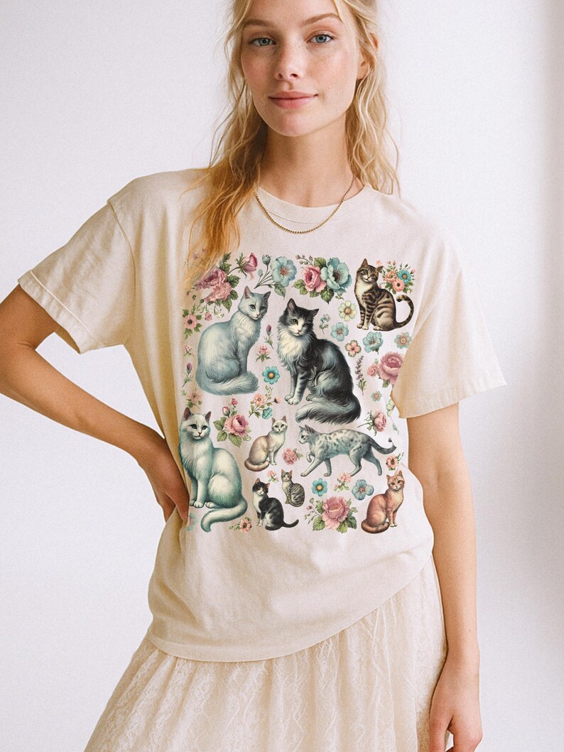 May include: A cream-colored short-sleeve t-shirt featuring a graphic print of multiple cats in various poses interspersed with colorful floral illustrations including pink and blue flowers across the front of the garment.