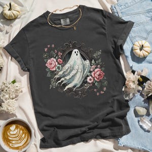 Puede incluir: Una camiseta gris oscuro con una ilustraci&oacute;n de estilo vintage de un fantasma en un marco floral. El fantasma es blanco y transl&uacute;cido, y las flores son rosas y moradas.