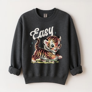 Puede incluir: Sudadera gris oscuro con una ilustraci&oacute;n de estilo vintage de un cachorro de tigre con rayas naranjas y negras. La palabra "Easy" est&aacute; escrita encima del tigre en escritura blanca. El dise&ntilde;o incluye peque&ntilde;os acentos de estrellas azules.