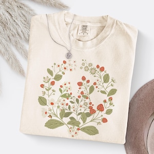 May include: A cream-colored t-shirt with a floral design featuring red strawberries and white flowers. The shirt has a crew neck and short sleeves. The text "COMFY COLORS" is printed on a small tag sewn into the shirt.