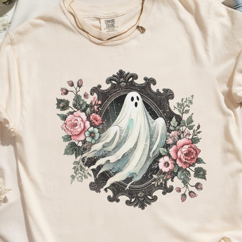 Puede incluir: Una camiseta blanca con un estampado gr&aacute;fico de un fantasma en un marco negro adornado rodeado de flores rosas y blancas.