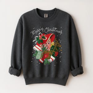May include: Dark gray sweatshirt with a festive Christmas design. The graphic features a vintage-style elf with a red hat and gifts. The words "Merry Christmas" are written in cursive above the elf. A cozy and seasonal apparel item.