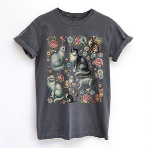 May include: A charcoal gray short-sleeve t-shirt featuring a central graphic print of multiple cats in various poses interspersed with colorful floral illustrations including pink, blue, and white flowers on the front of the garment.