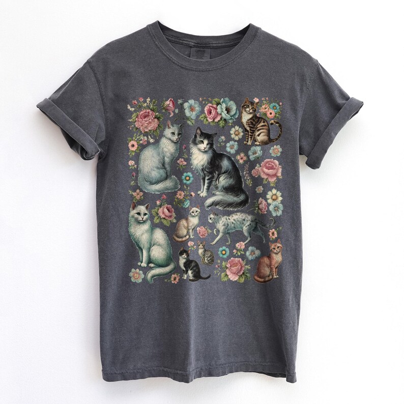 May include: A charcoal gray short-sleeve t-shirt featuring a central graphic print of multiple cats in various poses interspersed with colorful floral illustrations including pink, blue, and white flowers on the front of the garment.