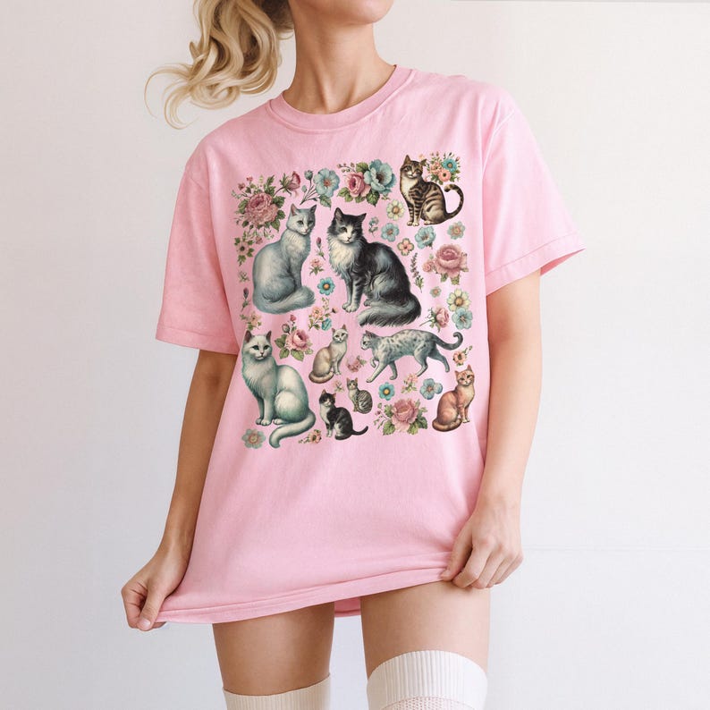 May include: A light pink short-sleeve t-shirt featuring a central graphic print of various cats in different poses and colors, interspersed with detailed illustrations of pink and blue flowers and green foliage across the front of the garment.