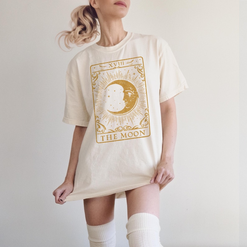 May include: A cream colored t-shirt with a gold tarot card design featuring the moon and the text "XVIII THE MOON".
