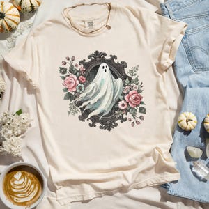 Puede incluir: Una camiseta blanca con una ilustraci&oacute;n de estilo vintage de un fantasma en un marco negro adornado rodeado de flores rosas y blancas.