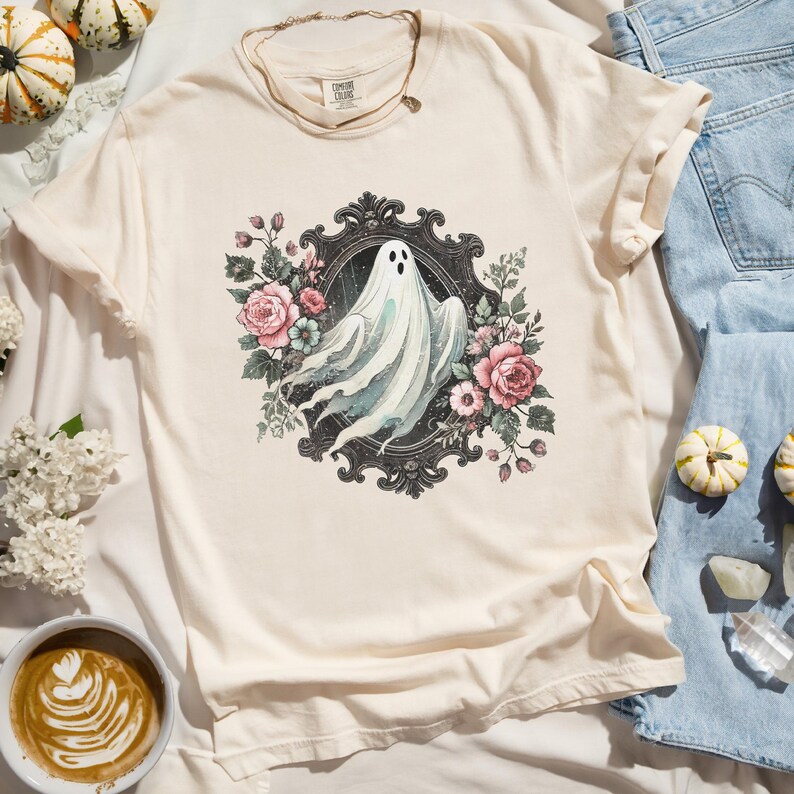 Puede incluir: Una camiseta blanca con una ilustraci&oacute;n de estilo vintage de un fantasma en un marco negro adornado rodeado de flores rosas y blancas.