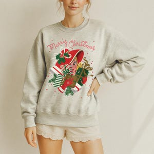 May include: A light gray sweatshirt with the words "Merry Christmas" in red script. The graphic features a woman in a red hat with gifts and greenery. The sweatshirt is a crew neck style.