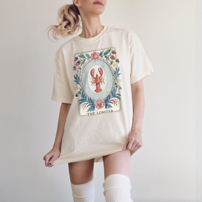 K&ouml;nnte beinhalten: Cremefarbenes T-Shirt mit einer detaillierten Illustration eines roten Hummers in einem floralen Rahmen. Das Design enth&auml;lt den Text "THE LOBSTER" am unteren Rand. Das Shirt hat kurze &Auml;rmel und eine lockere Passform, geeignet f&uuml;r Freizeitkleidung.