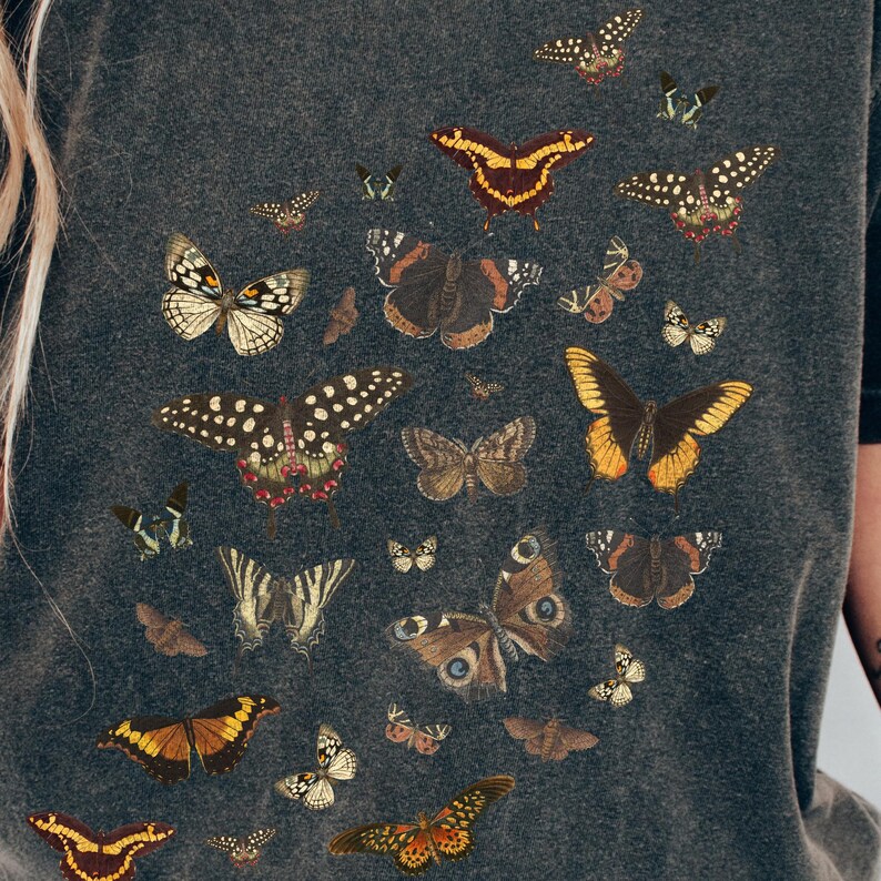 K&ouml;nnte beinhalten: Ein dunkelgraues T-Shirt mit einem Muster aus verschiedenen Schmetterlingen in unterschiedlichen Farben und Gr&ouml;&szlig;en.