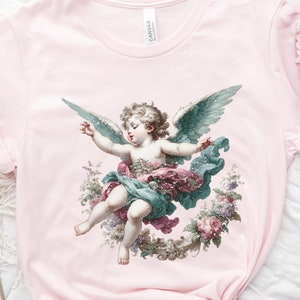 Puede incluir: Una camiseta rosa con una ilustraci&oacute;n de estilo vintage de un &aacute;ngel querub&iacute;n con alas, rodeado de flores. El &aacute;ngel lleva una t&uacute;nica azul y rosa y tiene una corona de flores en la cabeza.