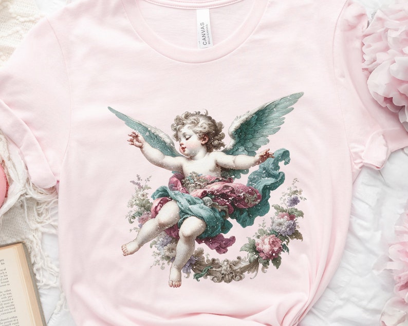 Puede incluir: Una camiseta rosa con una ilustraci&oacute;n de estilo vintage de un &aacute;ngel querub&iacute;n con alas, rodeado de flores. El &aacute;ngel lleva una t&uacute;nica azul y rosa y tiene una corona de flores en la cabeza.