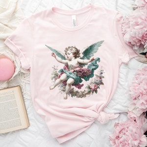 Puede incluir: Camiseta rosa con una ilustraci&oacute;n vintage de un &aacute;ngel querub&iacute;n con alas, rodeado de flores y vegetaci&oacute;n.