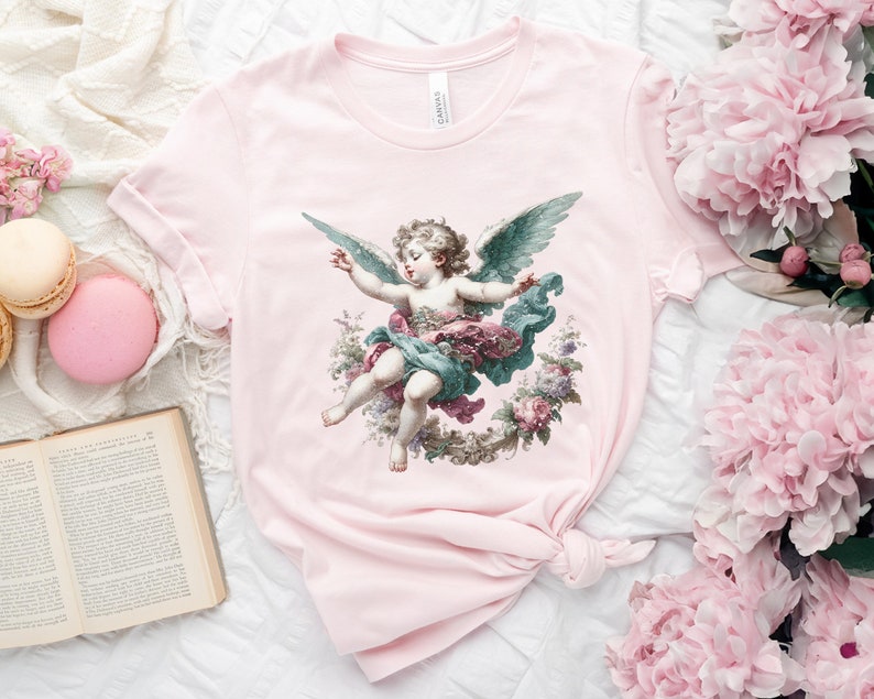 Puede incluir: Camiseta rosa con una ilustraci&oacute;n vintage de un &aacute;ngel querub&iacute;n con alas, rodeado de flores y vegetaci&oacute;n.