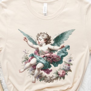 Puede incluir: Una camiseta de color crema con una ilustraci&oacute;n de estilo vintage de un &aacute;ngel querub&iacute;n con alas. El &aacute;ngel est&aacute; representado con una t&uacute;nica azul claro y verde, y el fondo presenta un dise&ntilde;o floral con flores rosas, blancas y verdes.