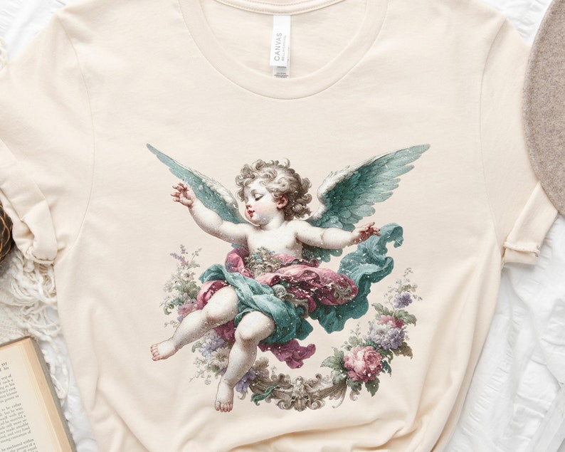 Puede incluir: Una camiseta de color crema con una ilustraci&oacute;n de estilo vintage de un &aacute;ngel querub&iacute;n con alas. El &aacute;ngel est&aacute; representado con una t&uacute;nica azul claro y verde, y el fondo presenta un dise&ntilde;o floral con flores rosas, blancas y verdes.