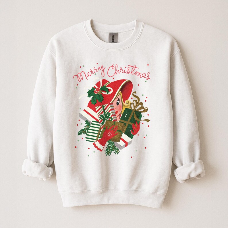 May include: White sweatshirt with a festive graphic. The graphic features a woman in a red hat holding gifts, with the words "Merry Christmas" in red script. The design includes green holly and red accents, perfect for the holiday season.
