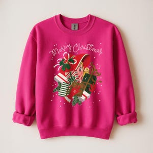 May include: A fuchsia sweatshirt with a festive Christmas design. The graphic features a woman in a red hat holding gifts, with the words "Merry Christmas" in cursive. White polka dots and green accents complete the holiday theme.