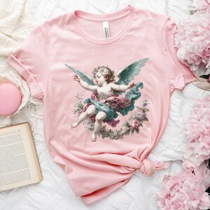 Puede incluir: Camiseta rosa con una ilustraci&oacute;n vintage de un &aacute;ngel querub&iacute;n con alas, rodeado de flores. El &aacute;ngel lleva una t&uacute;nica azul y verde y una corona de flores en la cabeza.