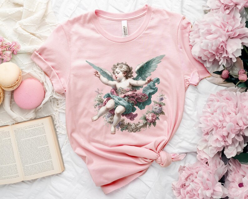 Puede incluir: Camiseta rosa con una ilustraci&oacute;n vintage de un &aacute;ngel querub&iacute;n con alas, rodeado de flores. El &aacute;ngel lleva una t&uacute;nica azul y verde y una corona de flores en la cabeza.