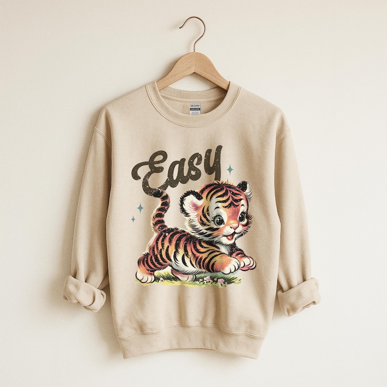 Puede incluir: Sudadera beige con una ilustraci&oacute;n de estilo vintage de un cachorro de tigre de dibujos animados. El tigre tiene rayas naranjas y negras y est&aacute; tumbado sobre hierba verde. La palabra "Easy" est&aacute; escrita encima del tigre.
