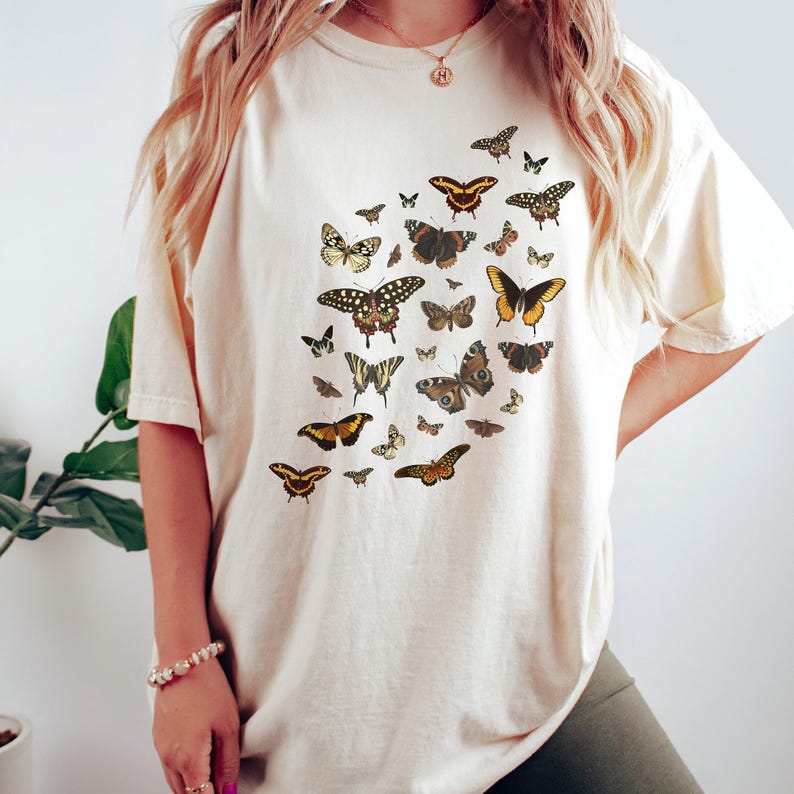 K&ouml;nnte beinhalten: Ein cremefarbenes T-Shirt mit einem grafischen Druck verschiedener Schmetterlinge in Braun-, Orange- und Gelbt&ouml;nen.