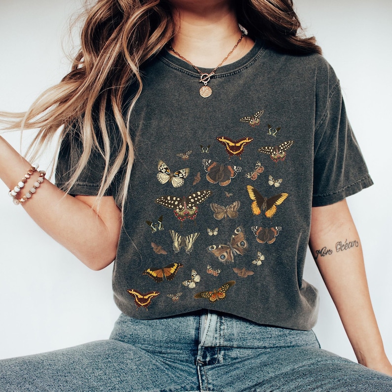 K&ouml;nnte beinhalten: Ein dunkelgraues T-Shirt mit einem Druck von verschiedenen Schmetterlingen in Braun-, Gelb- und Wei&szlig;t&ouml;nen. Die Schmetterlinge sind in einem zuf&auml;lligen Muster &uuml;ber das T-Shirt verteilt.