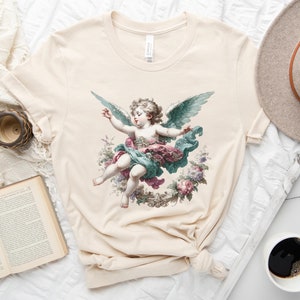 Puede incluir: Una camiseta beige con una ilustraci&oacute;n vintage de un querub&iacute;n con alas, rodeado de flores. El querub&iacute;n lleva un vestido azul y rosa.