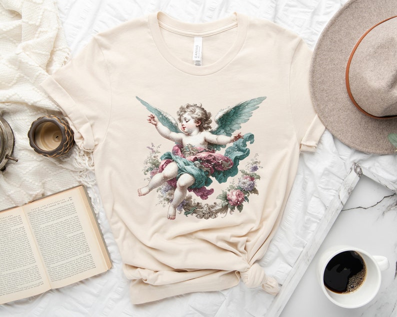 Puede incluir: Una camiseta beige con una ilustraci&oacute;n vintage de un querub&iacute;n con alas, rodeado de flores. El querub&iacute;n lleva un vestido azul y rosa.