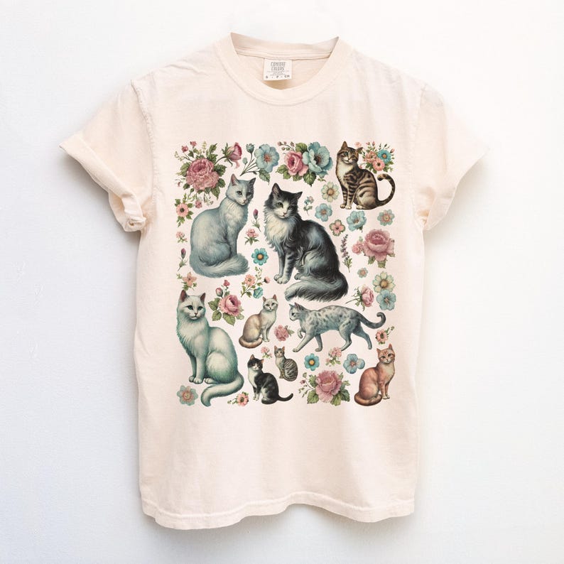 May include: A cream-colored short-sleeve t-shirt featuring a central graphic print of various cats in different poses interspersed with colorful floral illustrations including pink and blue flowers. The shirt has a crew neckline.