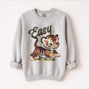 Puede incluir: Sudadera gris claro con una ilustraci&oacute;n de estilo vintage de un lindo cachorro de tigre con rayas negras y naranjas. La palabra "Easy" est&aacute; escrita encima del tigre en una fuente retro. Cuello redondo y mangas largas.