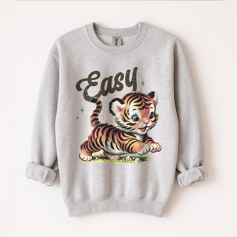 Puede incluir: Sudadera gris claro con una ilustraci&oacute;n de estilo vintage de un lindo cachorro de tigre con rayas negras y naranjas. La palabra "Easy" est&aacute; escrita encima del tigre en una fuente retro. Cuello redondo y mangas largas.