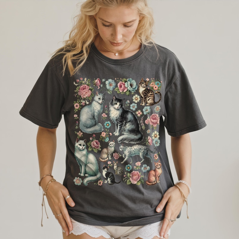 May include: A charcoal gray short-sleeve t-shirt featuring a central graphic print of multiple cats in various poses surrounded by colorful pink, blue, and white floral illustrations. The design includes a mix of tabby, solid, and spotted cats.