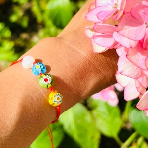 Può includere: Un braccialetto di corda rossa con perline colorate a forma di fiore. Le perline sono blu, verdi, gialle e bianche con una perla bianca.
