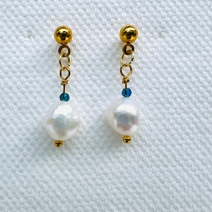 May include: A pair of gold-colored drop earrings with white baroque pearls. Each earring features a gold ball stud, a chain, a small blue bead, and a gold bead at the bottom. The earrings are displayed on a white textured surface.