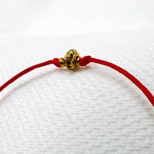 Può includere: Un braccialetto di corda rossa con un perno dorato. Il perno ha un design decorativo.