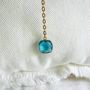 Op de afbeelding: Een goudkleurige ketting met een vierkante, lichtblauwe edelsteen hanger. De hanger is gezet in een gouden bezel en heeft een facetgeslepen oppervlak. De ketting wordt gepresenteerd op een witte, getextureerde ondergrond.