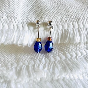 May include: A pair of dangle earrings featuring a silver ball stud, a thin silver bar, and a faceted, deep blue teardrop-shaped bead. The earrings also have a small gold and amber bead accent. The earrings are displayed on a white, textured surface.