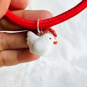 May include: A white chicken charm with a red comb and orange beak hangs from a silver ring on a bright red rope. The chicken charm is a small, detailed figurine. The rope is thick and textured, creating a bold contrast.