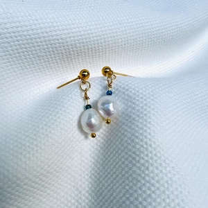 May include: A pair of gold earrings featuring white, oval pearls. Each earring has a small blue bead and a gold ball at the base. The earrings are displayed against a white, textured backdrop.