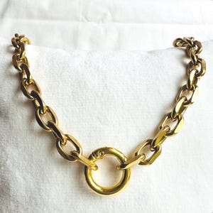 Peut inclure: Un collier en chaîne dorée avec un grand fermoir rond en or. La chaîne est composée de maillons épais de forme ovale.