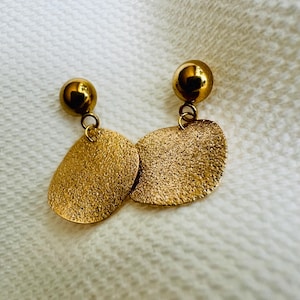 Op de afbeelding: Een paar goudkleurige oorbellen. Elke oorbel heeft een kleine, ronde stud verbonden met een getextureerde, ovaalvormige hanger. De oorbellen hebben een glanzende, metallic afwerking die licht reflecteert.