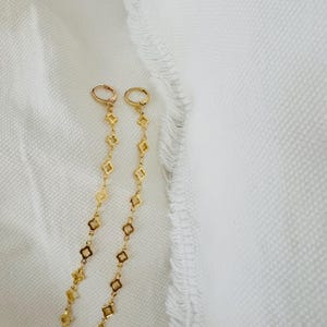 May include: A pair of gold-colored drop earrings. Each earring features a small hoop at the top, connected to a chain of diamond-shaped charms. The earrings are displayed against a white, textured background.