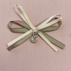 May include: A decorative bow made of three ribbons in beige, light brown, and off-white. A small, clear crystal charm hangs from the center of the bow. The bow is set against a pink fabric background.
