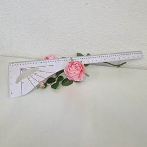 Könnte beinhalten: Ein weißes Plastiklineal mit einem Winkelmesser und Markierungen bis 127 cm. Das Lineal liegt auf einer weißen Oberfläche mit rosa Rosen.
