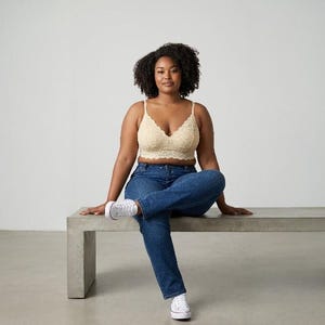 May include: A beige lace bralette with thin straps and a scalloped hemline paired with dark blue denim jeans and white sneakers. The bralette features a floral lace pattern throughout, while the jeans have a classic button and zipper fly closure.
