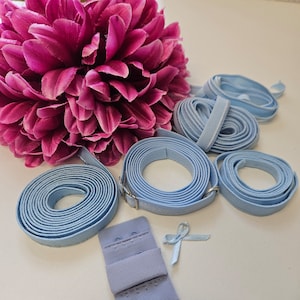 May include: A set of light blue elastic bra straps with metal clasps and a matching light blue bra strap adjuster. The straps are coiled and arranged on a white surface.