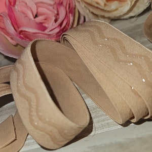 Könnte beinhalten: Beiges elastisches Band mit einem strukturierten, wellenförmigen Muster. Das Band ist gerollt und zeigt seine Breite und den subtilen Glanz des Materials. Weiche rosa und cremefarbene Blumen sind im Hintergrund.